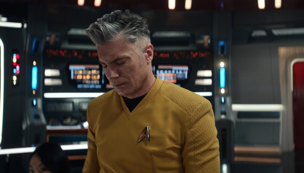 Image of Anson Mount as Captain Christopher Pike in a scene from 'Star Trek: Strange New Worlds.' He is a white man with perfectly coiffed salt-and-pepper hair wearing a gold Starfleet uniform. He's on the bridge of the Enterprise with his head bowed in frustration absolutely hating the fact that everyone has suddenly burst into song.