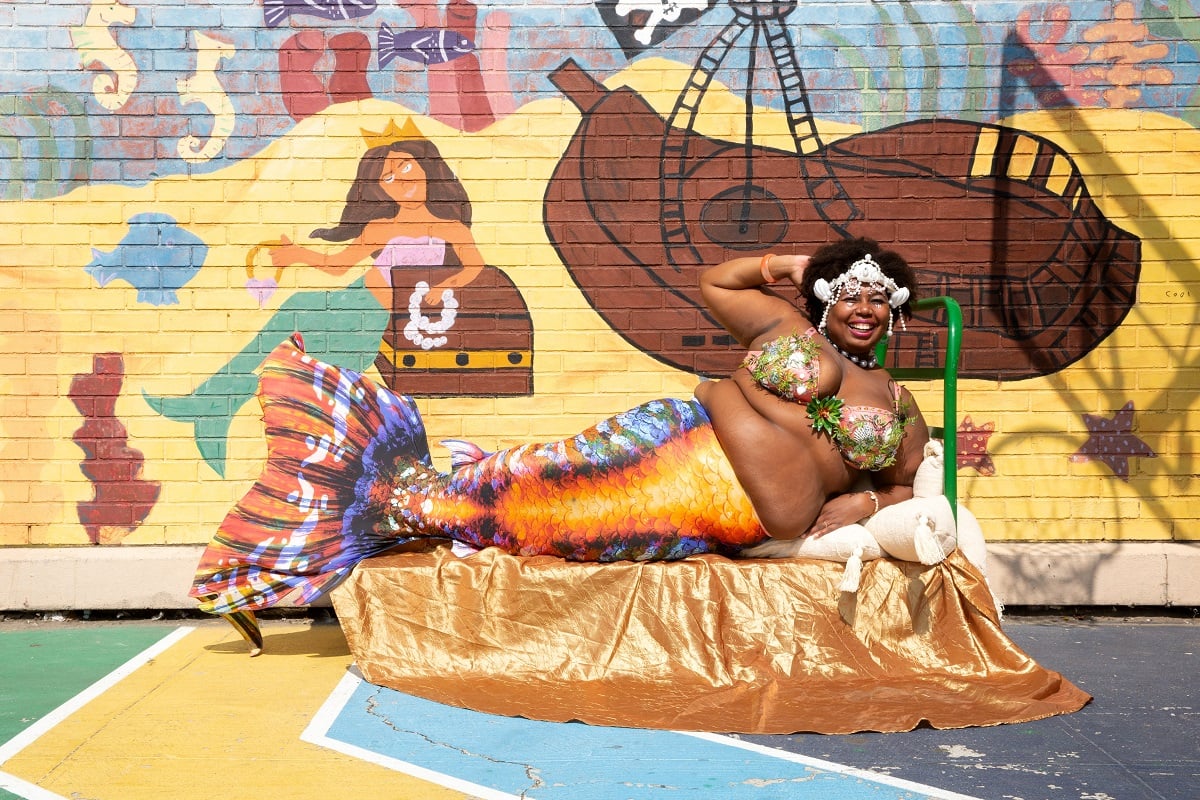 Image of a fat, Black woman smiling brightly as she poses while dressed as a mermaid in a scene from Netflix's 'MerPeople.' She has a tiara made of white seashells in her medium afro, a green floral bikini top, and an orange fish tail with blue accents. She's lying on a platform propped up on pillows in front of a mural on a brick wall depicting a mermaid and a crashed ship.