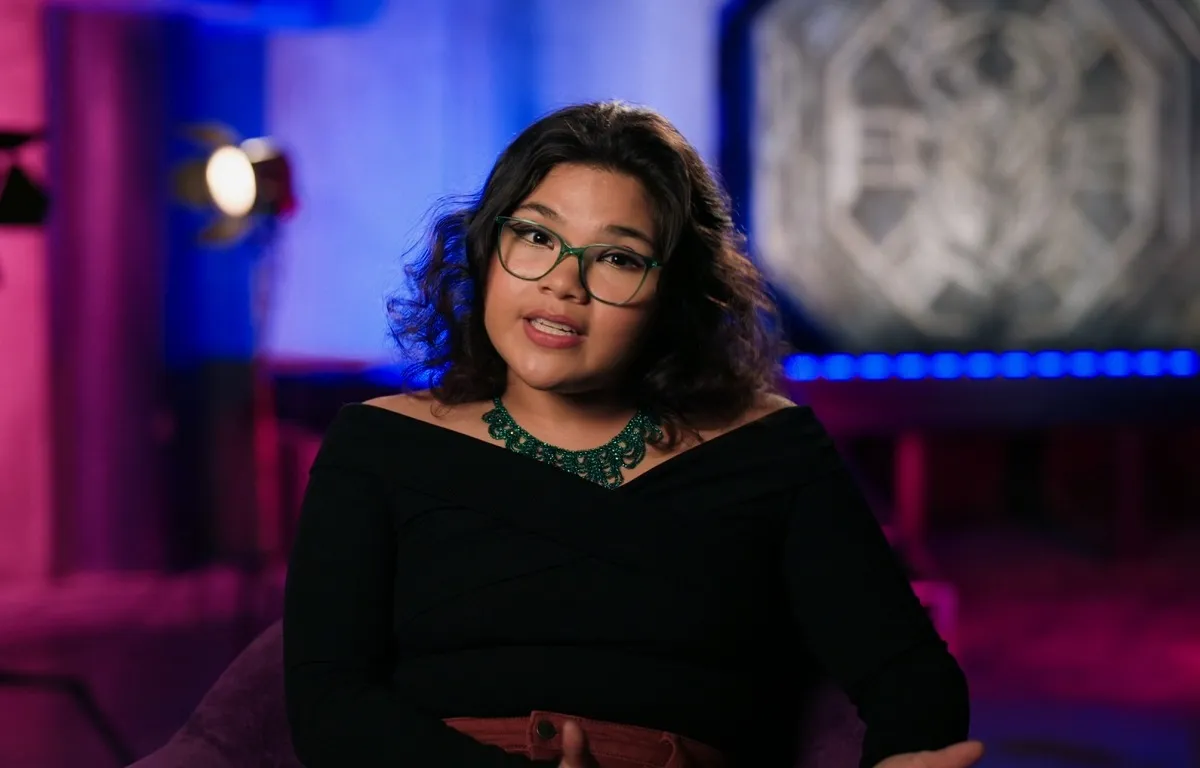 Screencap of Belissa Escobedo sitting on set in an EPK interview for 'Blue Beetle.' She is a brown Latina with thick, shoulder-length, wavy hair and is wearing green-rimmed glasses, a chunky green necklace, and a slightly off the shoulder, long-sleeved black shirt and a red belt.