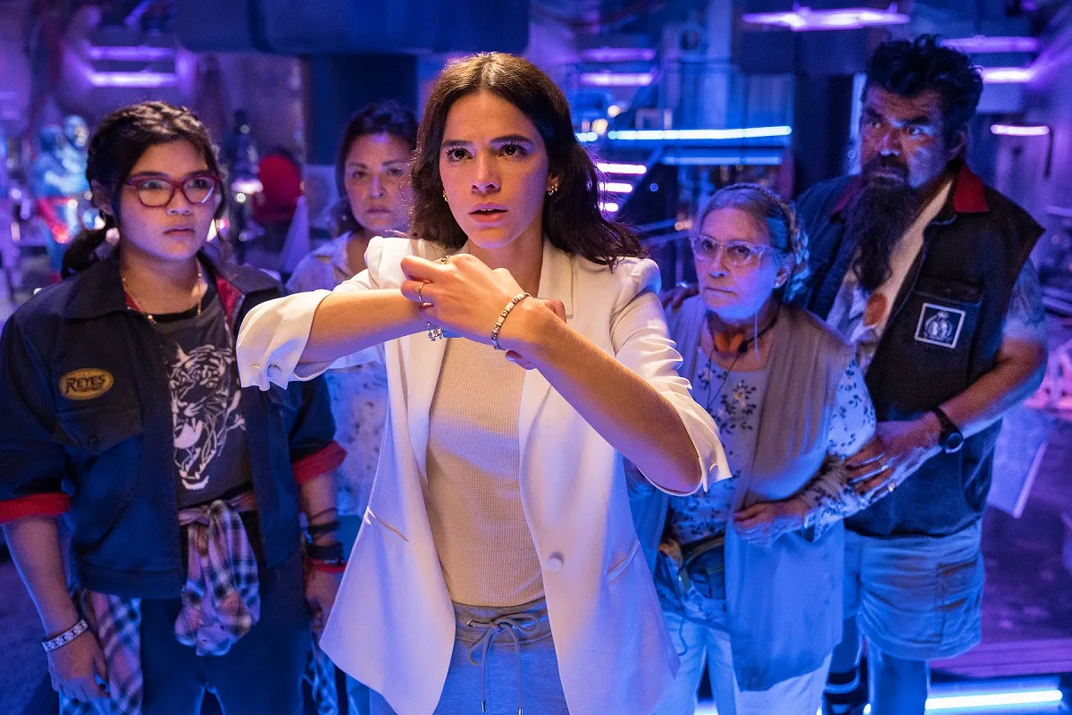 Milagro (Belissa Escobedo), Rocio (Elpidia Carrillo), Jenny (Bruna Marquezine), Nana (Adriana Barraza), and Uncle Rudy (George Lopez) in a scene from 'Blue Beetle.' Milagro, Rocio, Nana, and Rudy stand in a group behind Jenny, who is touching a device on her wrist.