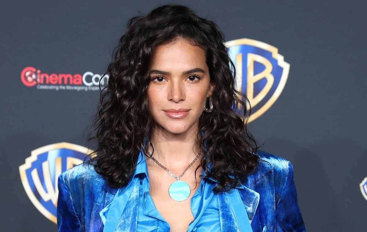 Publicity image of Bruna Marquezine for 'Blue Beetle.' She is standing in front of a black Warner Bros./CinemaCon step and repeat. She is a brown Brazililan with shoulder-length curly hair, and wearing thick silver triangular earrings, a necklace with a round, light blue pendant, and a velvet blue suit jacket with a lighter blue satin button-up shirt underneath. 