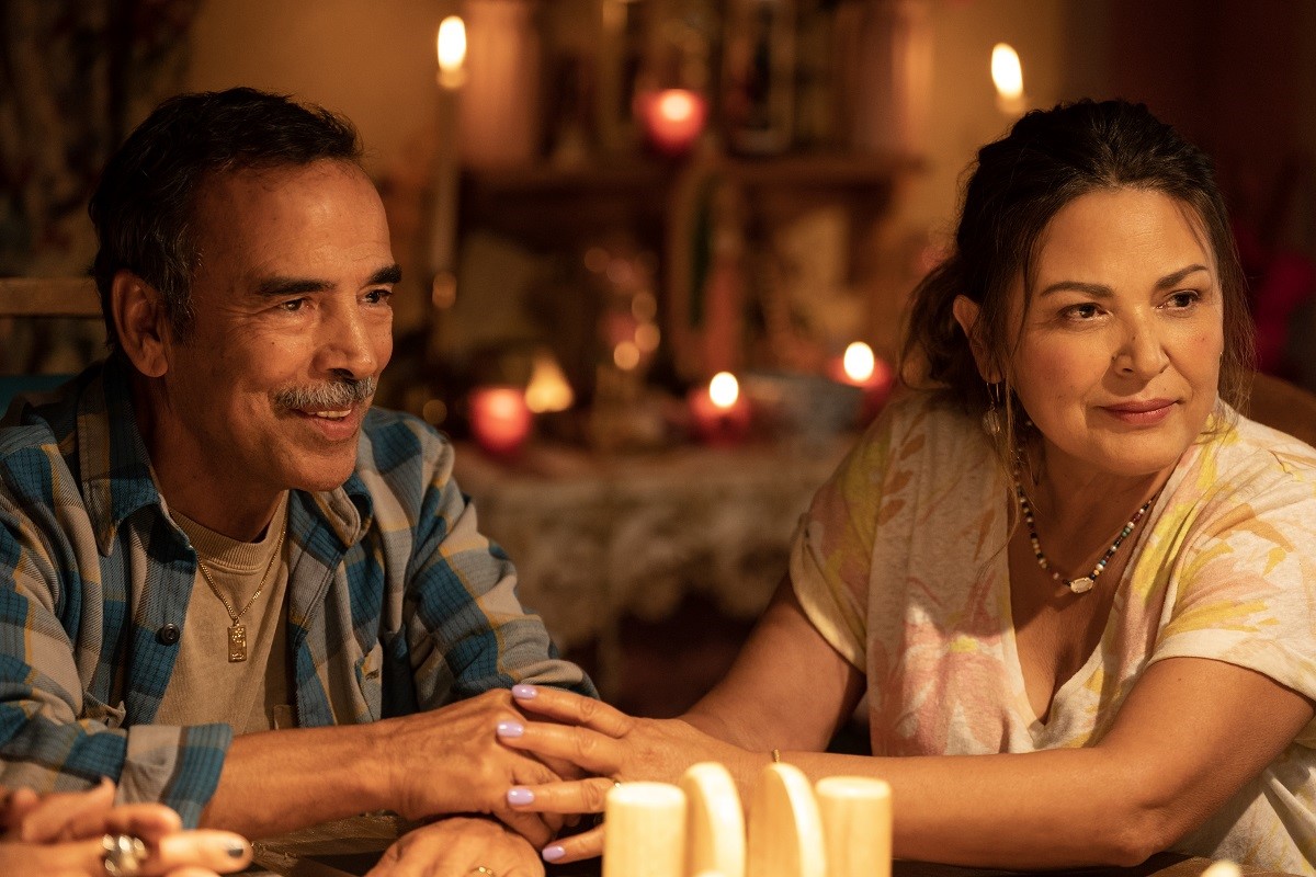 Damian Alcazar as Alfonso and Elpidia Carrillo as Rocio in a scene from 'Blue Beetle.' They are both middle-aged Latines and they're seated at a table holding hands as they look across the table at someone. Alfonso has close-cropped dark hair and a salt-and-pepper mustache and is wearing a blue plaid flannel shirt over a grey t-shirt. Rocio has long, dark hair pulled back into a ponytail, and she's wearing a blue and white beaded necklace and a white, orange, yellow, and pink floral print, v-neck short-sleeved shirt. 