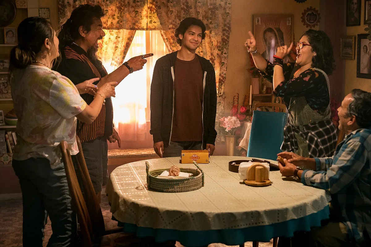 ELPIDIA CARRILLO as Rocio, GEORGE LOPEZ as Uncle Rudy, XOLO MARIDUEÑA as Jaime Reyes, BELISSA ESCOBEDO as Milagro and DAMIAN ALCAZAR as Alberto in a scene from 'Blue Beetle.' They are all brown Latines, and everyone is standing around a dining room table laughing about something, except for Alberto, who is seated. They're all making a joke at Jaime's expense as he smiles sheepishly.