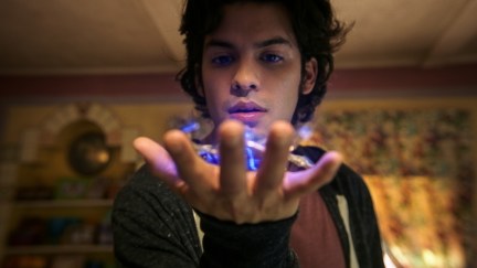 Xolo Maridueña as Jaime Reyes in a scene from 'Blue Beetle.' He is a brown Latino with shaggy dark hair wearing a black hoodie over a red t-shirt. He holds his hand outstretched holding a glowing blue scarab.