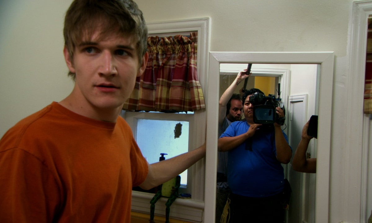 Image of Bo Burnham as Zach Stone in a scene from 'Zach Stone is Gonna Be Famous.' He is a white teenage boy with shaggy blondish hair and wearing an orange t-shirt. He's holding up a full-length mirror in his kitchen to reveal a camera crew in the doorway - a fat, brown woman holding the camera, and a white man behind her holding a boom mic. 