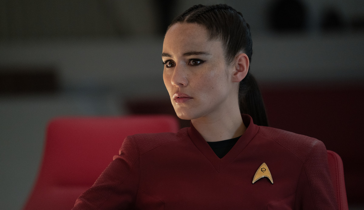 Image of Christina Chong as La'an Noonien-Singh in a scene from 'Star Trek: Strange New Worlds." She is a mixed race white and Chinese woman with dark hair pulled tightly back in a ponytail formed by two braids. She's wearing a red Starfleet uniform as she sits seriously at a conference table.