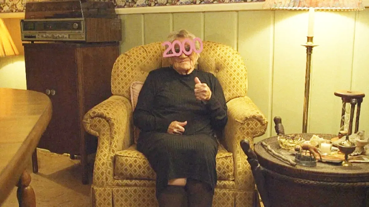 An old woman (Coe-Jane Weight) giving the camera a thumbs up while wearing party glasses in V/H/S/99