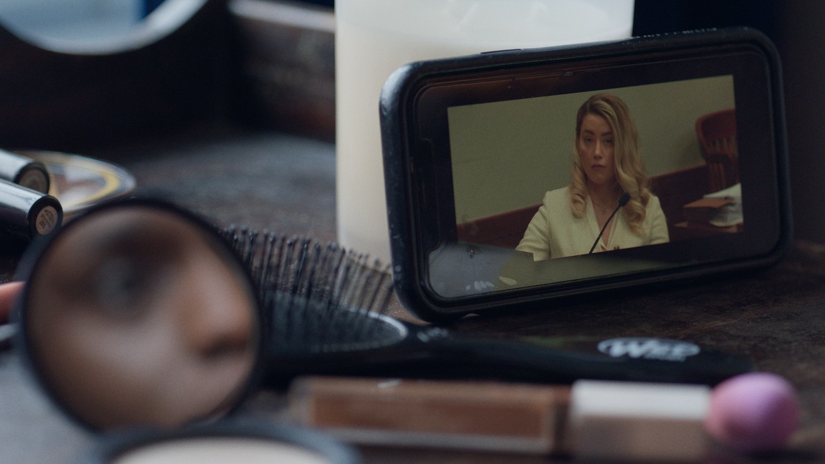 Netflix's 'Depp v Heard': A woman, seen in a compact mirror reflection, is watching Amber Heard give testimony on a phone screen while doing her makeup.