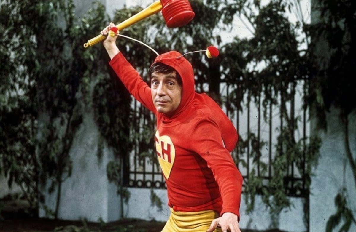 Promotional image of Chespirito in costume as the title character on El Chapulín Colorado. He is a white Latino with short, dark hair wearing a red, long-sleeved hoodie with long, grasshopper-like antennae on the hood. There's a big, yellow heart in the center of the shirt that has the letters "CH" in red on it. He's wearing yellow pants, and holding up a red and yellow plastic sledgehammer. 