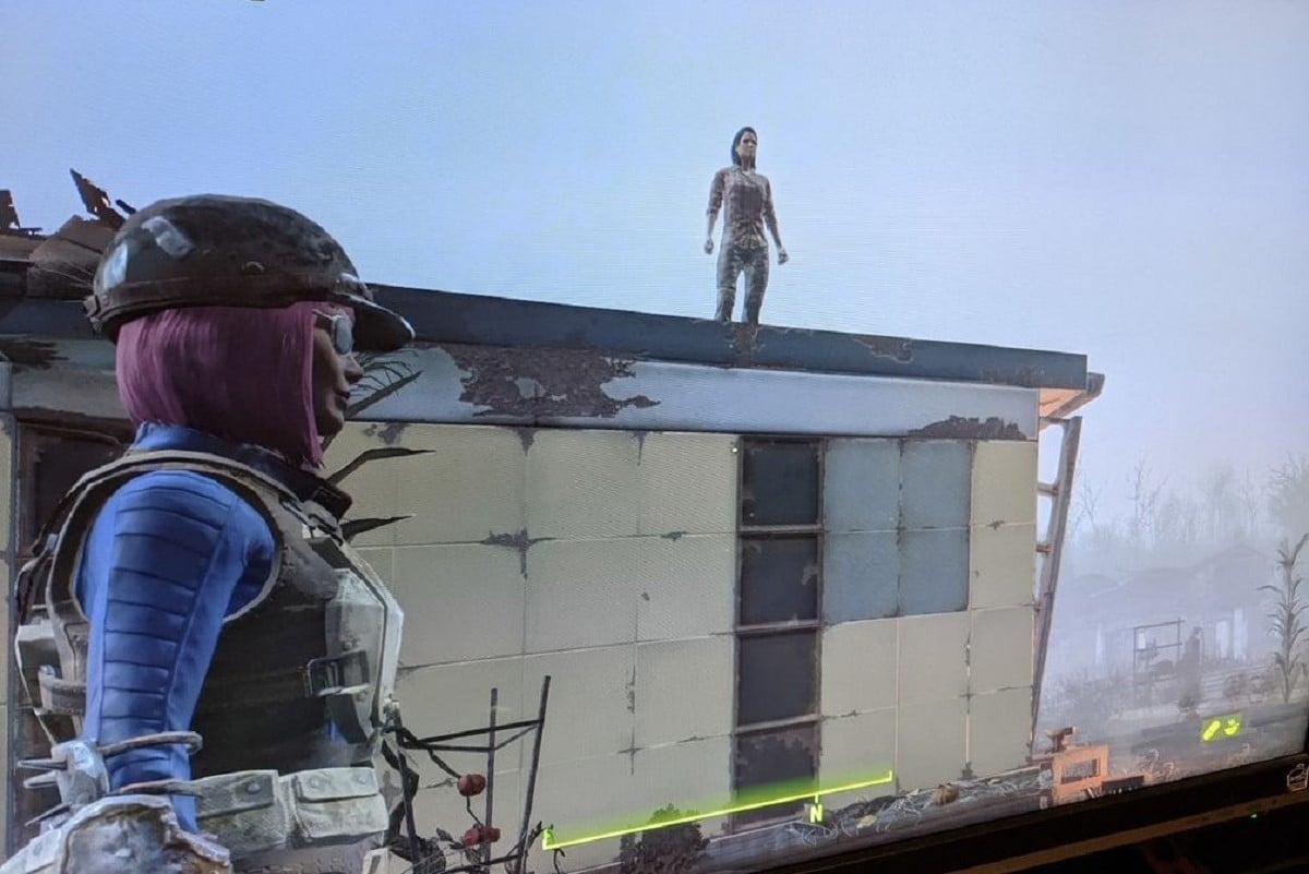 Screencap from the game 'Fallout 4.' Teresa Jusino's female player character is seen in profile wearing her blue Vault suit under body armor while looking at a house where the NPC Marcy Long is standing on the roof.