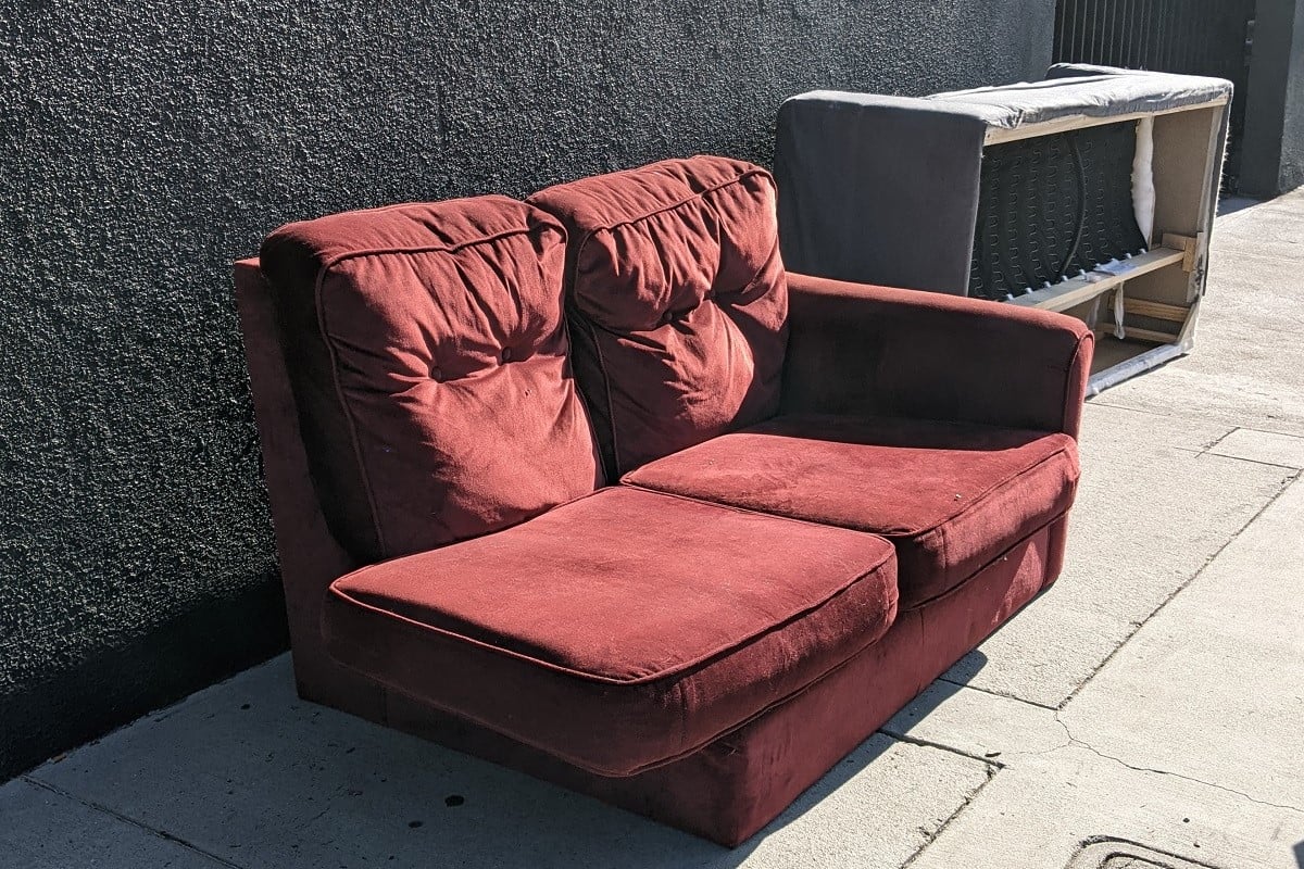 Photograph of two couches left out on the sidewalk as trash. In the foreground is a red fabric couch that looks like one part of a sectional. Behind it is a grey couch that's been flipped over onto its back.