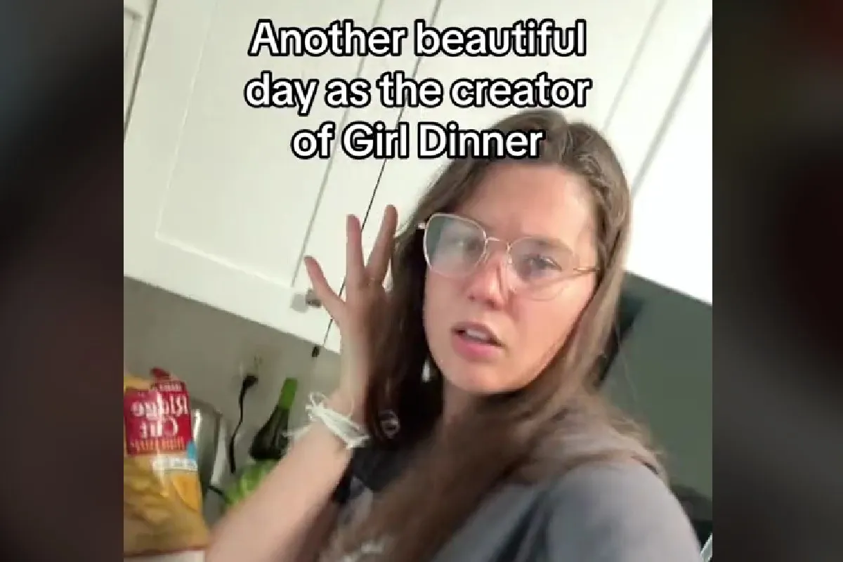 Screencap of a TikTok featuring @liviemaher. She is a young, white woman with long, brown hair wearing thin-rimmed glasses and a grey t-shirt. She's holding a hand to her ear as she holds her phone up in her other hand to shoot a video as she stands in her kitchen. The text above her reads "Another beautiful day as the creator of Girl Dinner."