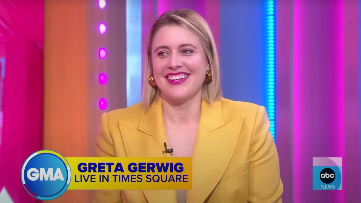 Greta Gerwig doing an interview on ABC's 'Good Morning America.' She is a white woman with shoulder-length blonde hair wearing a yellow suit jacket and thick gold hoop earrings. She's smiling and sitting in front of a brightly lit pink/blue/purple/orange backdrop. The chyron on the screen reads "GMA: Greta Gerwig - Live in Times Square" and there's an ABC News logo in the lower right hand corner.