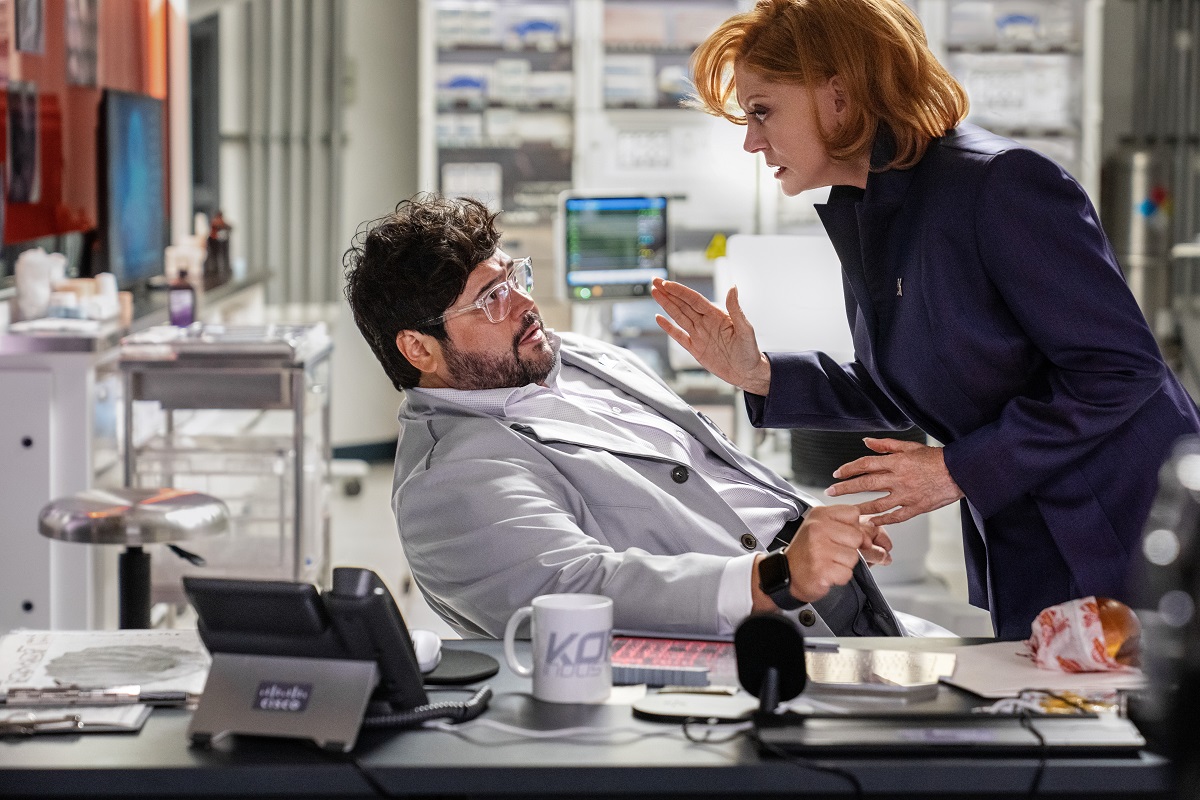 Victoria Kord (Susan Sarandon) and "Dr. Sanchez" (Harvey Guillen) in a scene from 'Blue Beetle.' Victoria is leaning threateningly over Sanchez, who is sitting in a desk chair and leaning back fearfully. 