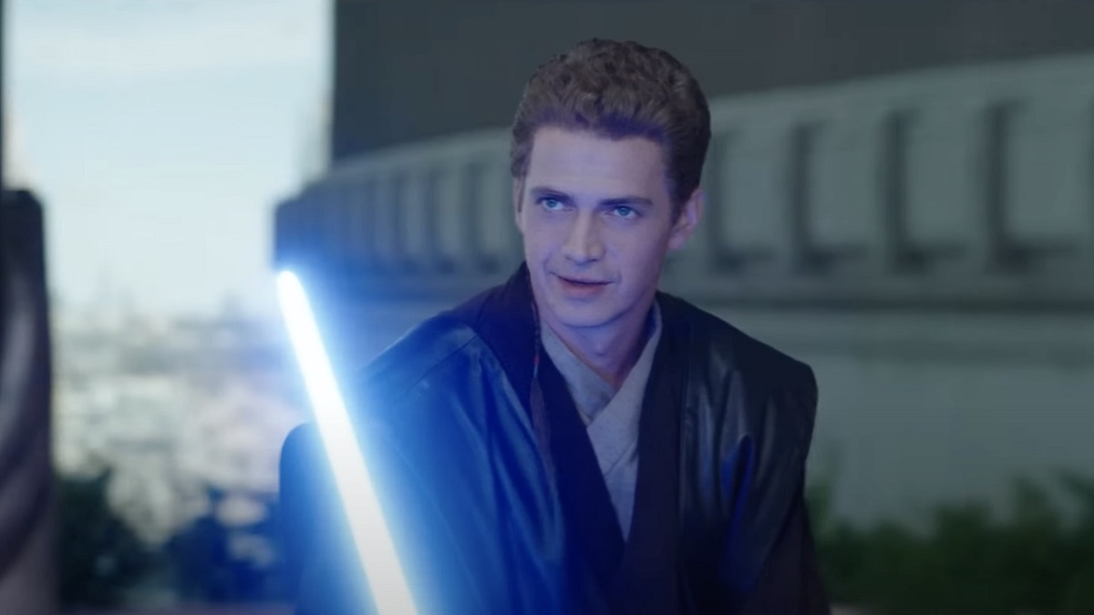 Hayden Christensen as Anakin Skywalker in a scene from 'Obi-Wan Kenobi.' He is a white man with short, sandy brown hair. He's wearing a black leather cloak over beige Jedi robes and is holding a blue light saber as he looks amusedly at someone across from him.