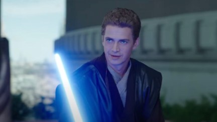 Hayden Christensen as Anakin Skywalker in a scene from 'Obi-Wan Kenobi.' He is a white man with short, sandy brown hair. He's wearing a black leather cloak over beige Jedi robes and is holding a blue light saber as he looks amusedly at someone across from him.