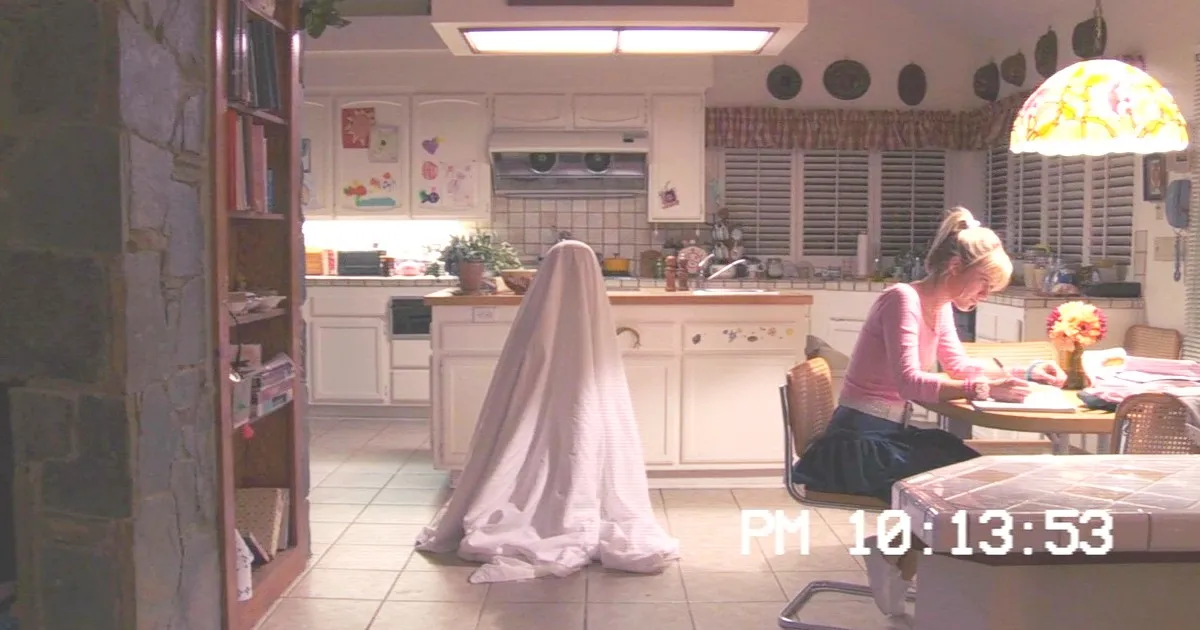 Lisa (Johanna Braddy) sitting in the kitchen, unaware of the entity under a sheet behind her in Paranormal Activity 3