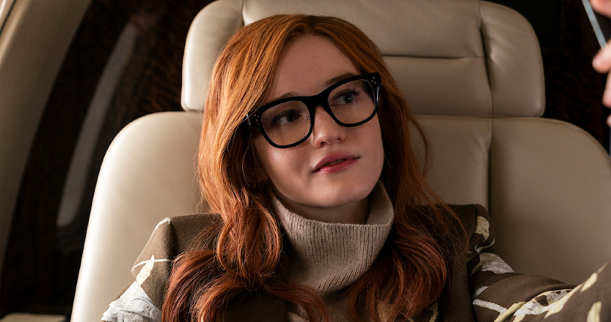 Julia Garner as Anna Sorokin/Anna Delvey in 'Inventing Anna.' She is a stylish young woman wearing glasses, seated in a leather chair