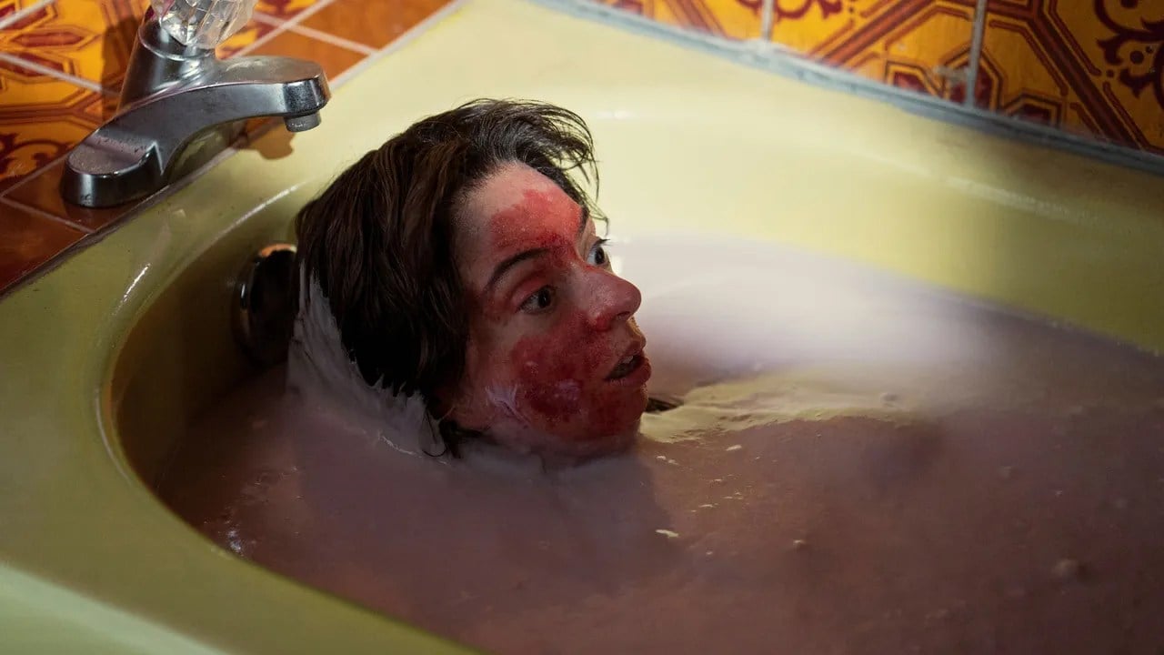 A woman with her face covered in blood sits in a bath of cloudy water.