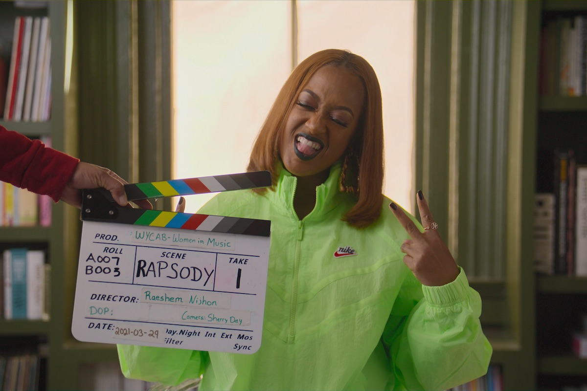 Rapper Rapsody throws a peace sign while a hand holds a filming slate in front of her face before filming a documentary scene.
