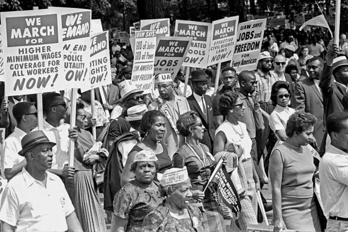The March on Washington - Marchers gathering at the Lincoln Memorial after walking from Washington Monument grounds. The image depicts a crowd of people marching, holding up placard that read "WE MARCH FOR HIGHER MINIMUM WAGES / COVERAGE FOR ALL WORKERS NOW" and similar.