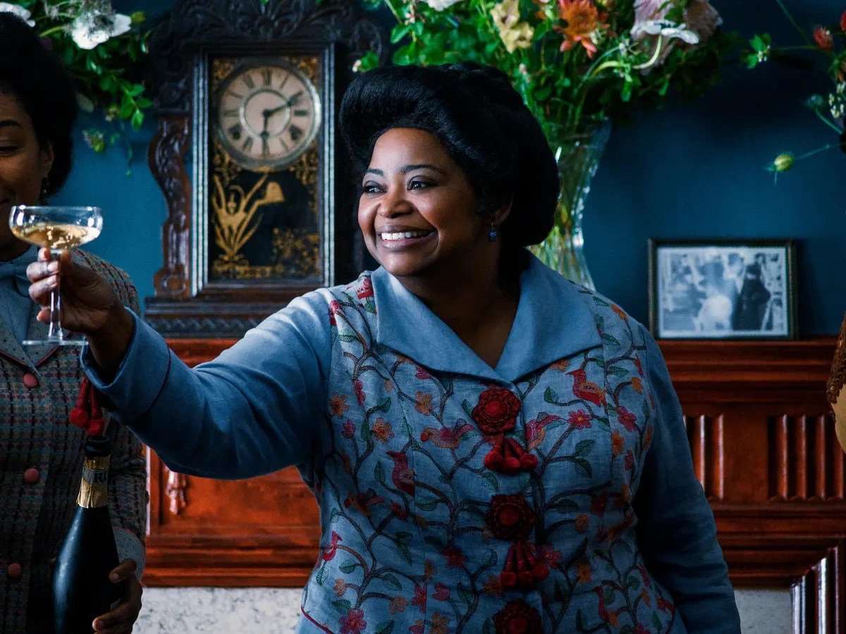 Image of Octavia Spencer as Sarah Breedlove in a scene from Netflix's 'Self-Made.' She is a Black woman dressed in a 19th century blue dress with floral embroidery on it and large red buttons with tassels. Her black hair is pulled up into an updo and she smiles as she toasts a glass of champagne.
