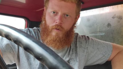 Oliver Anthony, a young white man with a red beard, sits in a truck.