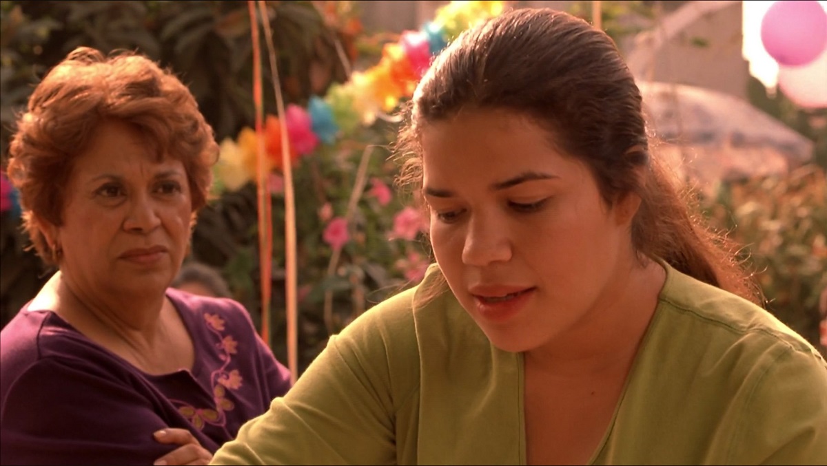 Lupe Ontiveros as Carmen and America Ferrera as Ana in 'Real Women Have Curves.' Ana is a Latina high school senior. Her long, dark hair is in a ponytail, and she's wearing a green, v-necked long-sleeved shirt as she looks down at something she's doing. Carmen is an older Latina with short, reddish, curly hair wearing a long-sleeved, purple shirt with pink floral embroidery at the collar. Her arms are folded as she looks at Ana sternly.