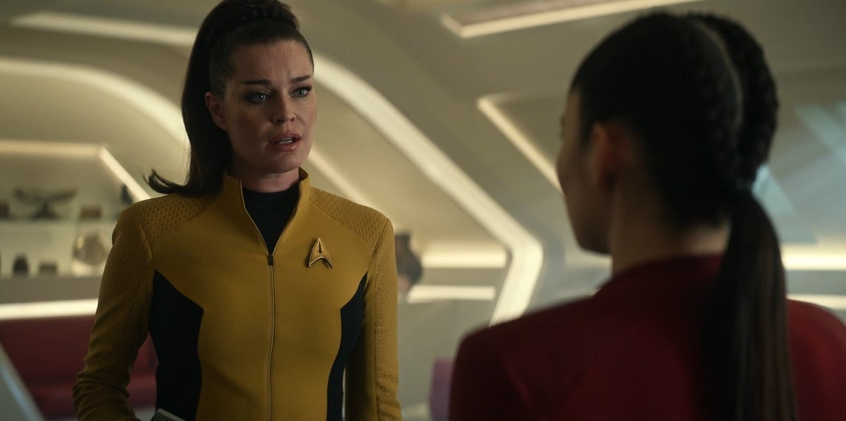 Image of Rebecca Romijn as Una Chin-Riley in a scene from 'Star Trek: Strange New Worlds." She is a white woman with long dark hair pulled into a high, 1960s-style ponytail and wearing a gold Starfleet uniform. She is singing to La'an who stands out of focus in the foreground with her back to the camera.