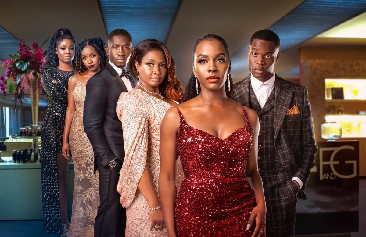 The all-Black cast of the Amazon Prime show, 'Riches.' The six cast members are standing in an office setting wearing upscale dresses or suits with Deborah Ayorinde standing in the foreground wearing a red sequined sleeveless dress with a low neckline, dangly diamond earrings, and her long, black hair slicked back in an elegant ponytail.