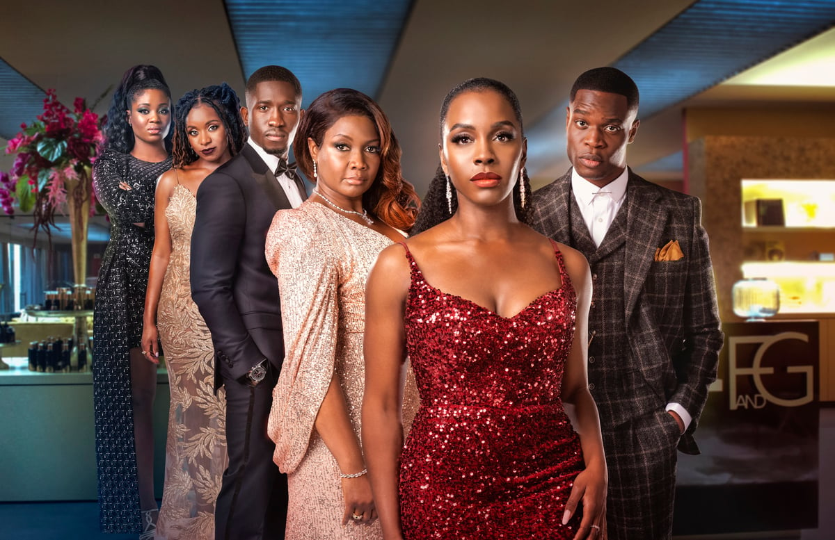 The all-Black cast of the Amazon Prime show, 'Riches.' The six cast members are standing in an office setting wearing upscale dresses or suits with Deborah Ayorinde standing in the foreground wearing a red sequined sleeveless dress with a low neckline, dangly diamond earrings, and her long, black hair slicked back in an elegant ponytail. 