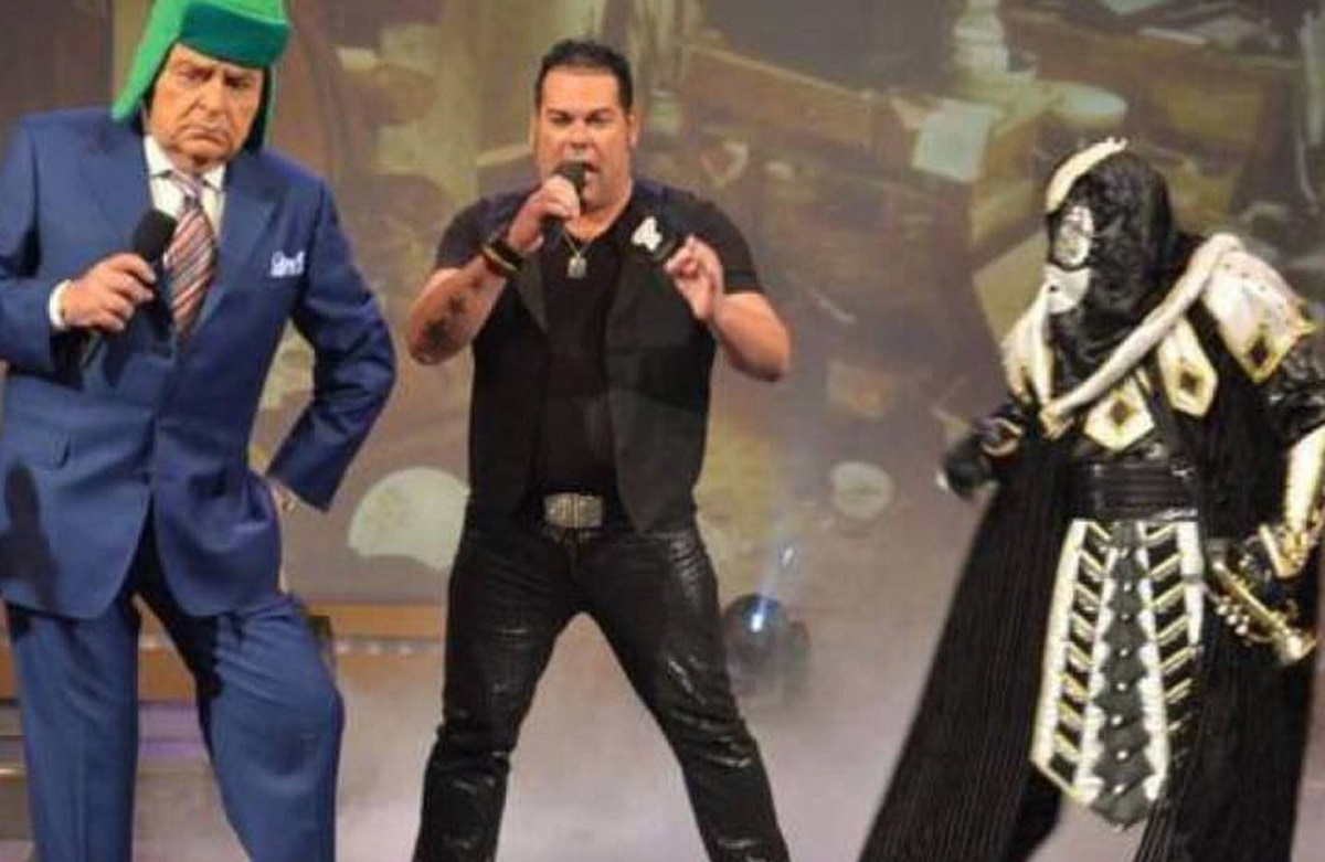 Image from the show 'Sabado Gigante.' Host Don Francisco is on the left, and is an older, light-skinned Latino in a blue suit and wearing a green ushanka hat while dancing and holding a microphone. In the center is a light-skinned Latino wearing a black t-shirt, black vest, and black leather pants singing into the microphone. On the right is the character El Chacal, a masked character in an all-black costume with white accents holding a trumpet. 