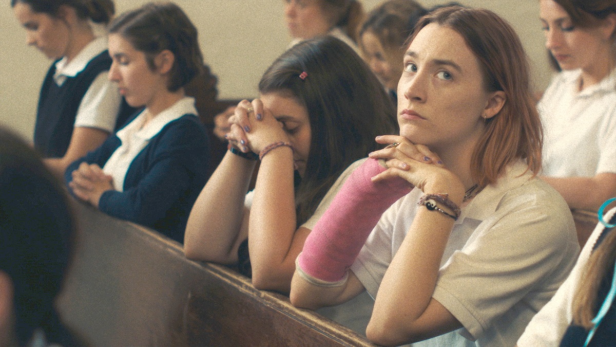 Saiorse Ronan as the titular character in 'Lady Bird.' She is a white teenage girl with shoulder-length blond hair wearing a white, short-sleeved polo shirt and a pink cast on one arm as she kneels in a church pew surrounded by other girls, her hands folded as she looks off to the side, bored.