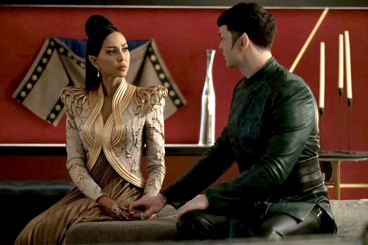 Gia Sandhu as T'Pring and Ethan Peck as Spock in a scene from 'Star Trek: Strange New Worlds.' T'Pring has brown skin, and long dark hair that's styled half up in a bun. She is a Vulcan with pointed ears and is wearing an elaborately adorned white jacket with gold trim over a pleated gold dress. Spock is white with short dark hair, also pointed ears, wearing a dark green long-sleeved top and black pants. They are seated on a bed, and Spock is reaching out holding T'Pring's hand.