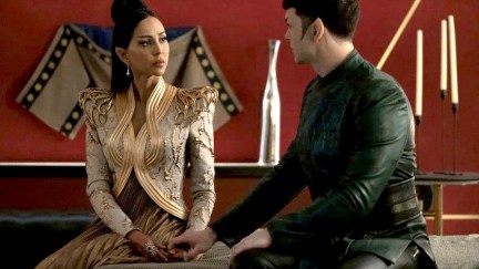 Gia Sandhu as T'Pring and Ethan Peck as Spock in a scene from 'Star Trek: Strange New Worlds.' T'Pring has brown skin, and long dark hair that's styled half up in a bun. She is a Vulcan with pointed ears and is wearing an elaborately adorned white jacket with gold trim over a pleated gold dress. Spock is white with short dark hair, also pointed ears, wearing a dark green long-sleeved top and black pants. They are seated on a bed, and Spock is reaching out holding T'Pring's hand.