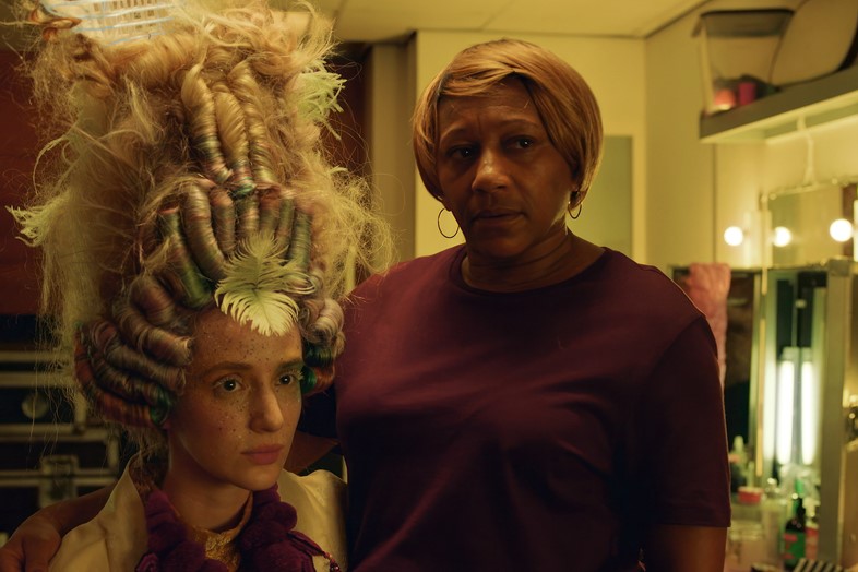 A model sits with a huge powdered wig hairstyle, with her stylist next to her.