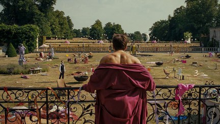 A white man with his robe draped to his shoulders looks at mess on the lawn on a palatial estate in 