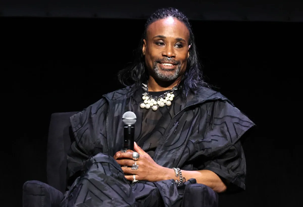 Billy Porter, sitting and holding a microphone, looking to the side and smiling.