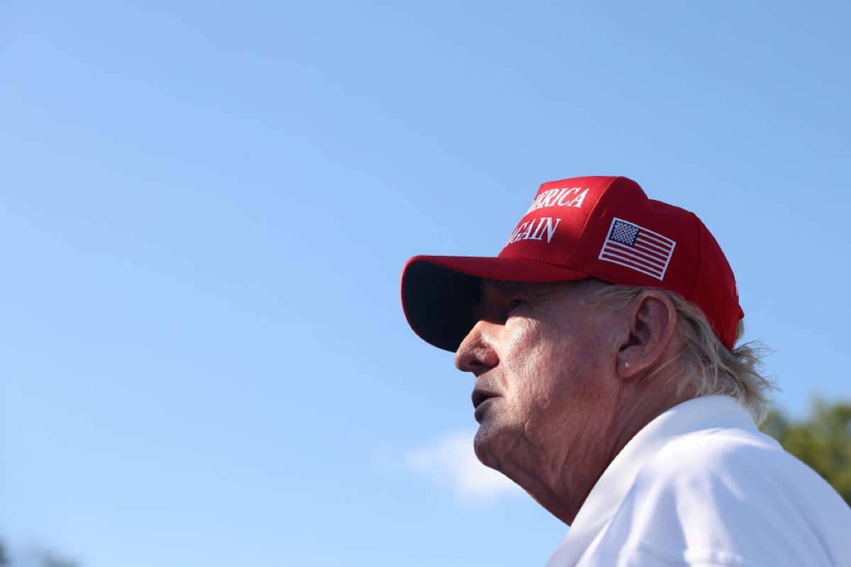 Donald Trump looking into the distance, against a blue sky background, wearing his fascist hat.
