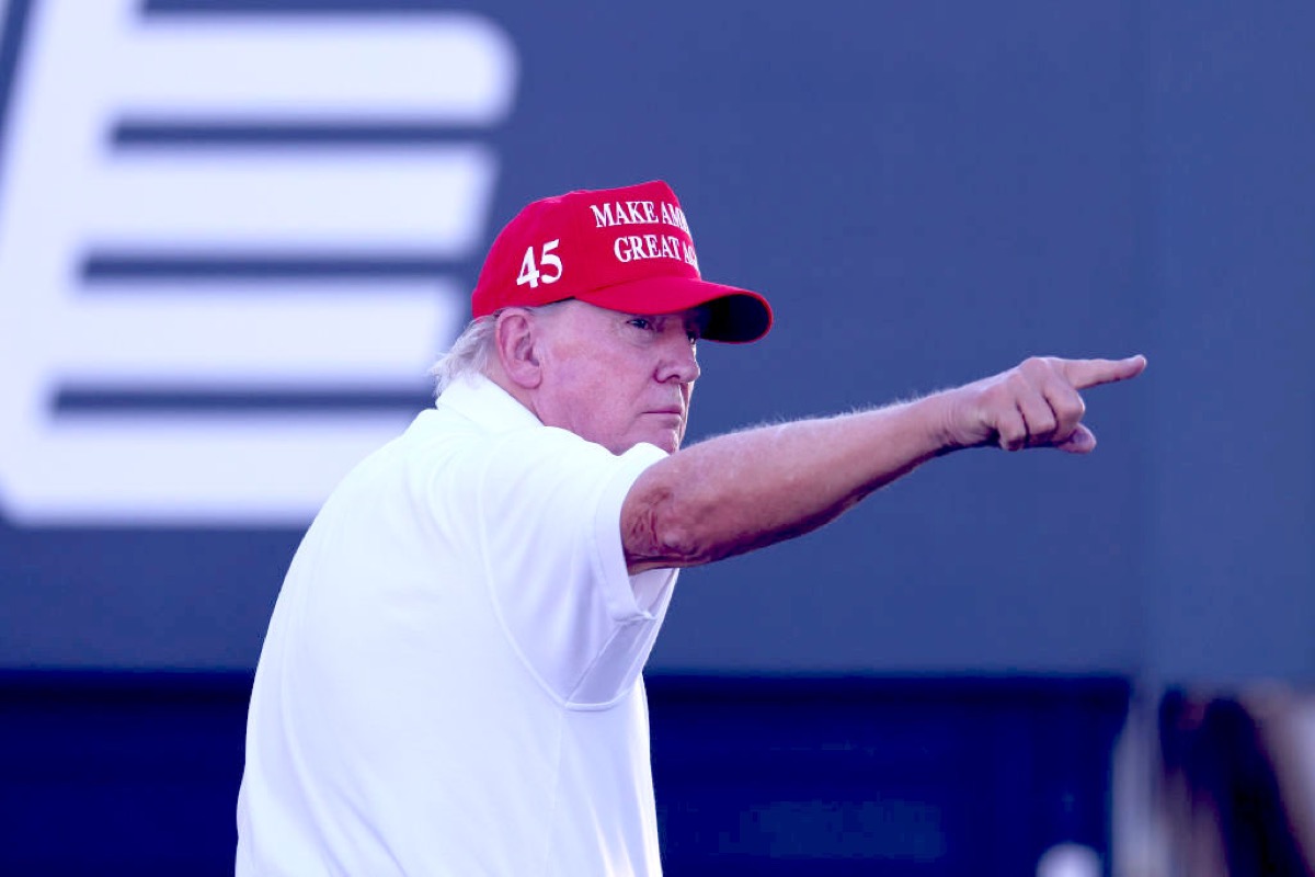 Donald Trump, looking off to the side and pointing, wearing his fascist hat.