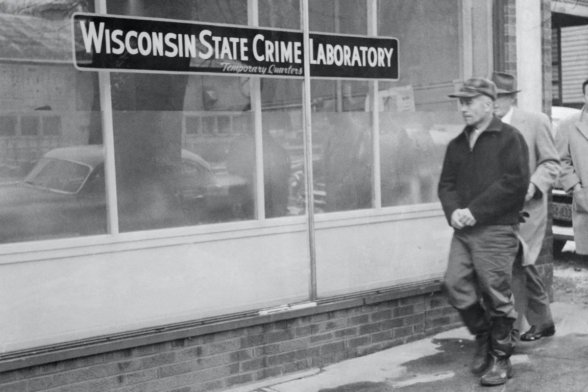 A black and white photograph of Ed Gein walking by a sign reading Wisconsin State Crime Laboratory