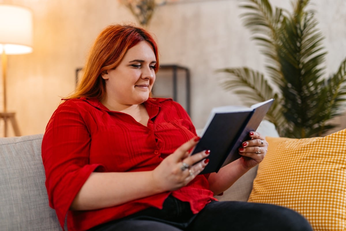 A fat woman reads a book while sitting on her couch.