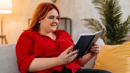 A fat woman reads a book while sitting on her couch.