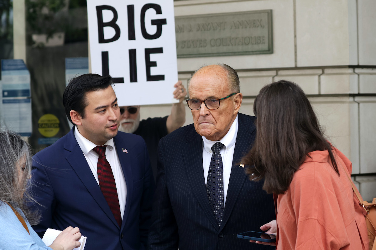 Rudy Giuliani talking to the press outside a courthouse. A protester stands behind him with a large sign reading "BIG LIE"