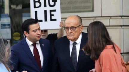 Rudy Giuliani talking to the press outside a courthouse. A protester stands behind him with a large sign reading 