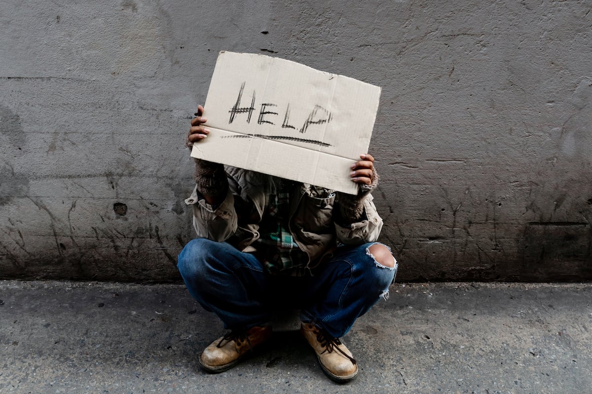 A homeless man sits hopelessly leaning against a wall as there is no one to help him with work and food in his hand holding a sign for help.