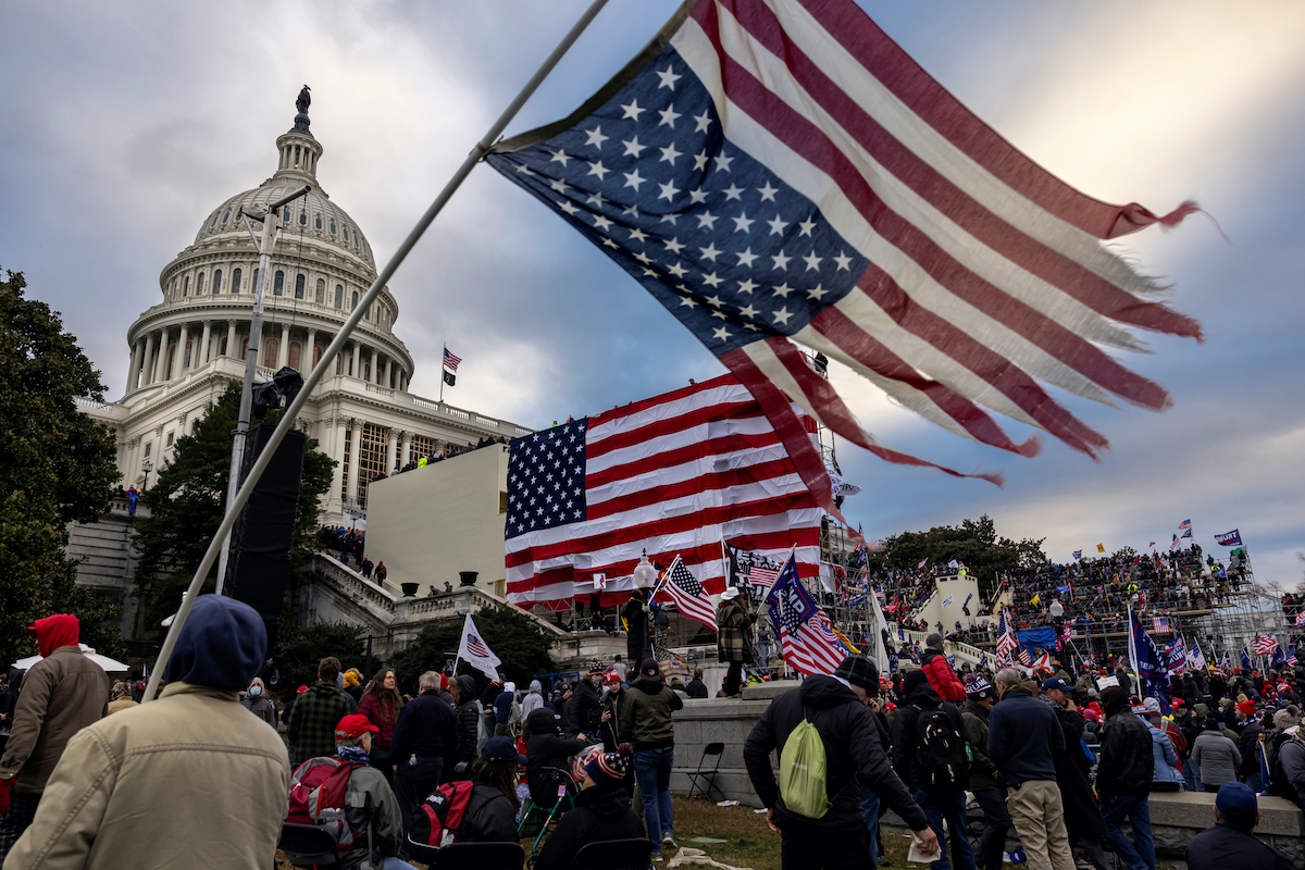 The US Capitol surrouded by a mob of Trump supporters on January 6. A large shredded American flag is being waved in the foreground.