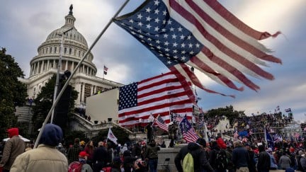 The US Capitol surrouded by a mob of Trump supporters on January 6. A large shredded American flag is being waved in the foreground.