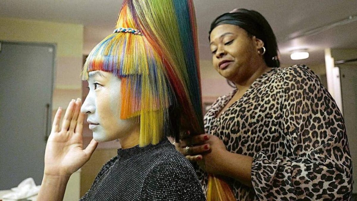 A model with her face painted white and tall, rainbow-colored hair sits in a chair with a stylist standing behind her.