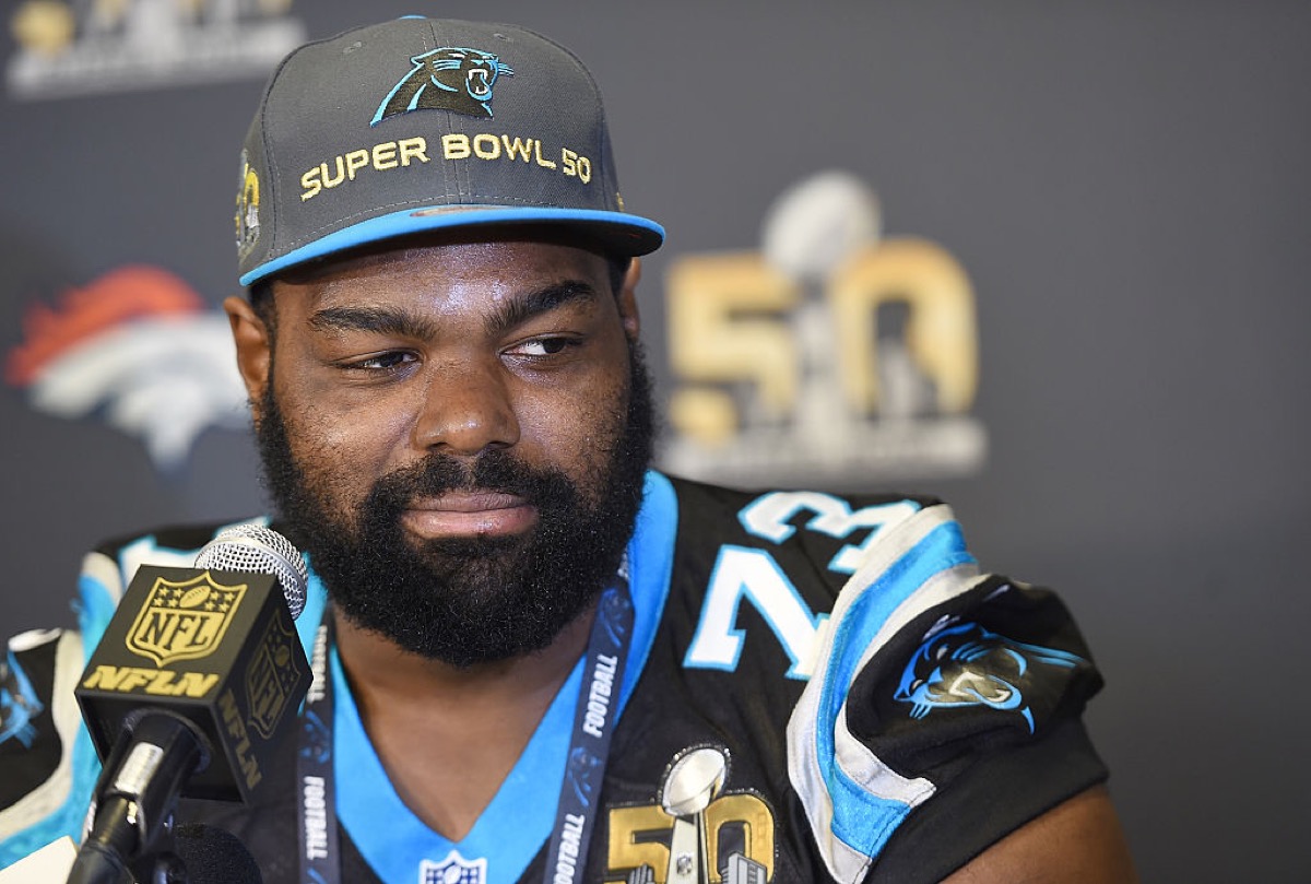 Michael Oher sits at a microphone in his football uniform at a press conference, looking to the side skeptically.